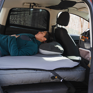 Person lying down on a makeshift bed in the back of a car. The back seats are removed, creating space for a mattress and pillow. Various camping gear is seen organized in the front seat and hanging on the back of the driver's seat.