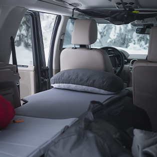Interior of a vehicle set up for camping, with a bed and pillow extending from the backseat. Outside, snowy trees are visible through the open door and windows, and there are bags and camping gear around the bed.