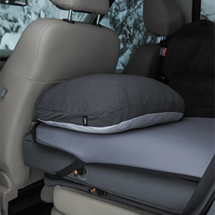 A gray pillow rests on a folded mattress in the backseat of a car. The car's interior includes beige seats and a headrest. Snowy outdoor scenery can be seen through the window in the background.