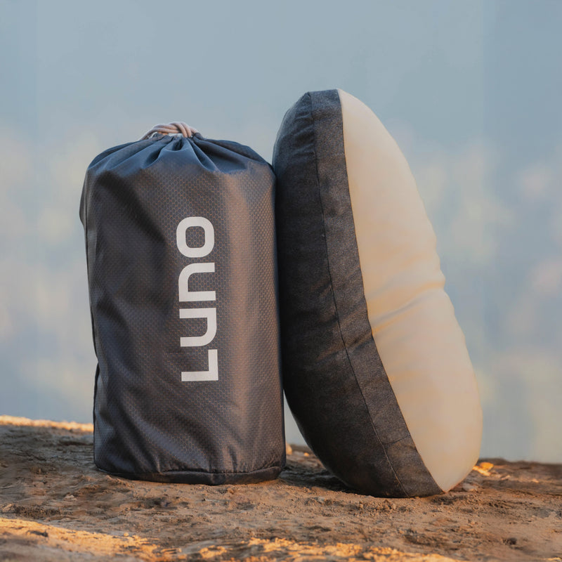 A Luno® Truck Camping AIR Starter Bundle, featuring a portable pillow in a black case labeled "Luno" and the upright pillow beside it on dirt, is the ideal car camping essential for restful sleep in nature.