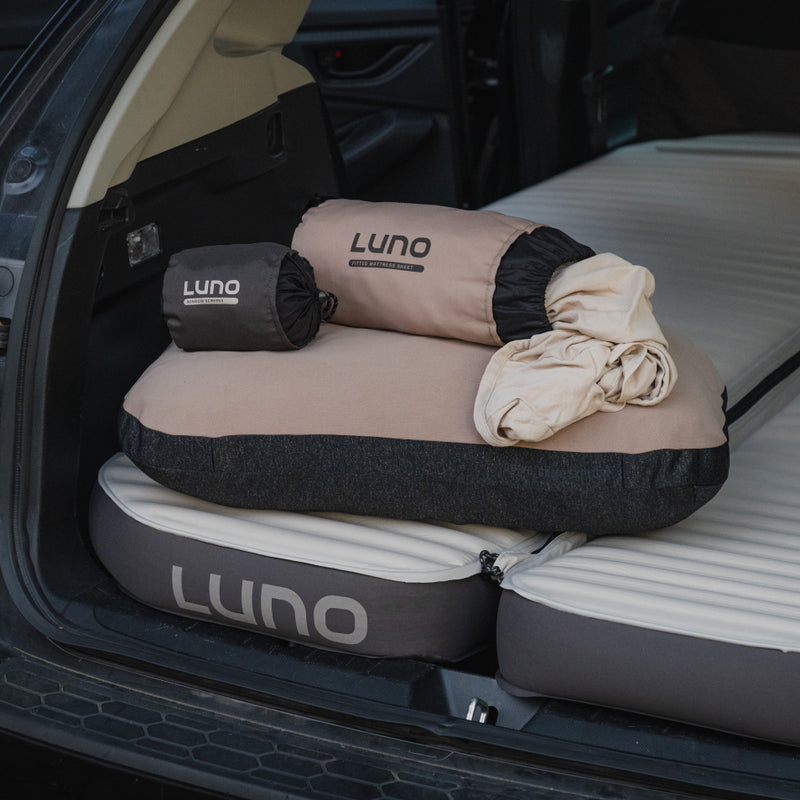 The Luno® Car Camping AIR+FOAM Pro Starter Bundle—including a travel pillow, rolled blanket, and storage bags—is displayed on an inflated car mattress in the open back of a vehicle, ideal for comfortable outdoor adventures.
