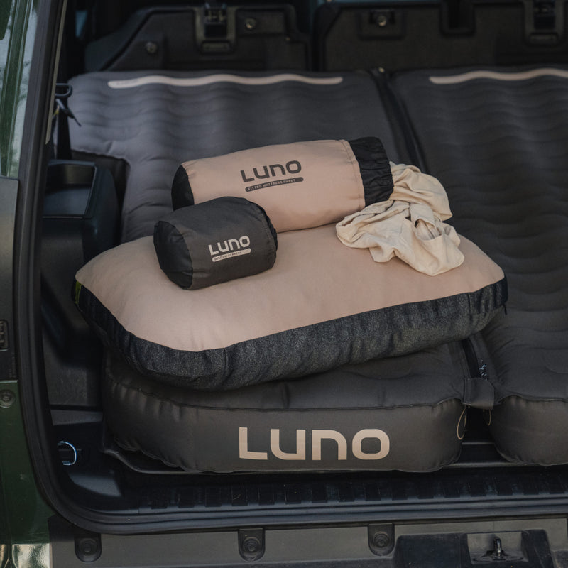 A vehicle trunk displays the Luno® Car Camping AIR Pro Starter Bundle—an inflatable mattress, pillow, and two compact storage bags—showcasing essentials for a comfortable car camping or road trip adventure.