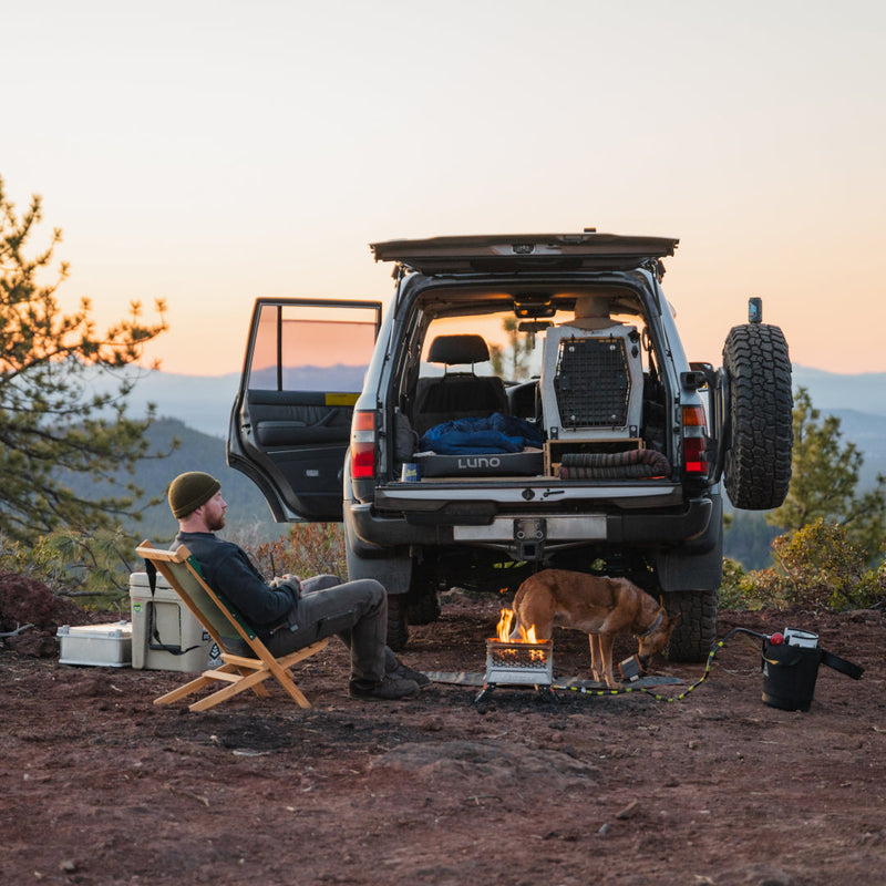 A person wearing a beanie relaxes in a camping chair next to a parked SUV on a hillside, where a campfire is burning and a dog is nearby. The SUV's back is open, revealing an AIR+FOAM PRO Camping Mattress by Luno HQ, which appears in like-new condition. The scenic backdrop includes trees and distant mountains under a clear sky.