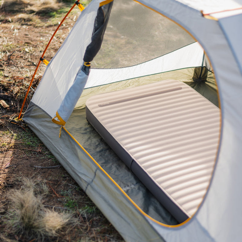 A camping tent is pitched on dry ground, featuring an AIR+FOAM PRO Camping Mattress by Luno HQ in a like-new condition inside. The tent is partially open, revealing its mesh sides and orange poles. Dry grass and scattered leaves can be seen surrounding the tent.