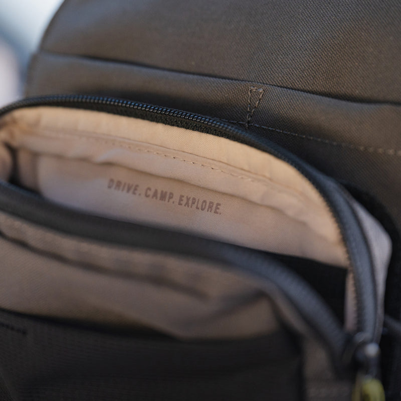 A close-up shot of an open pocket on the dark-colored Headrest Organizer by Luno HQ reveals the words "DRIVE. CAMP. EXPLORE." elegantly stitched into the fabric lining inside, showcasing its practical organization with multiple compartments for all your essentials.