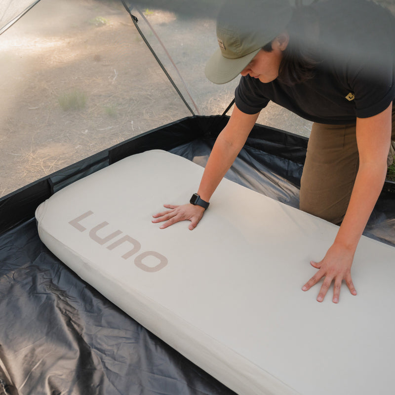 A person sets up an inflatable mattress labeled "Luno®" inside a tent. Wearing a green cap and black shirt, they lean forward to adjust the Fitted Mattress Sheet - AIR+FOAM Pro Camping Mattress, which is placed on the black tent floor. Natural light filters into the tent, illuminating the scene.