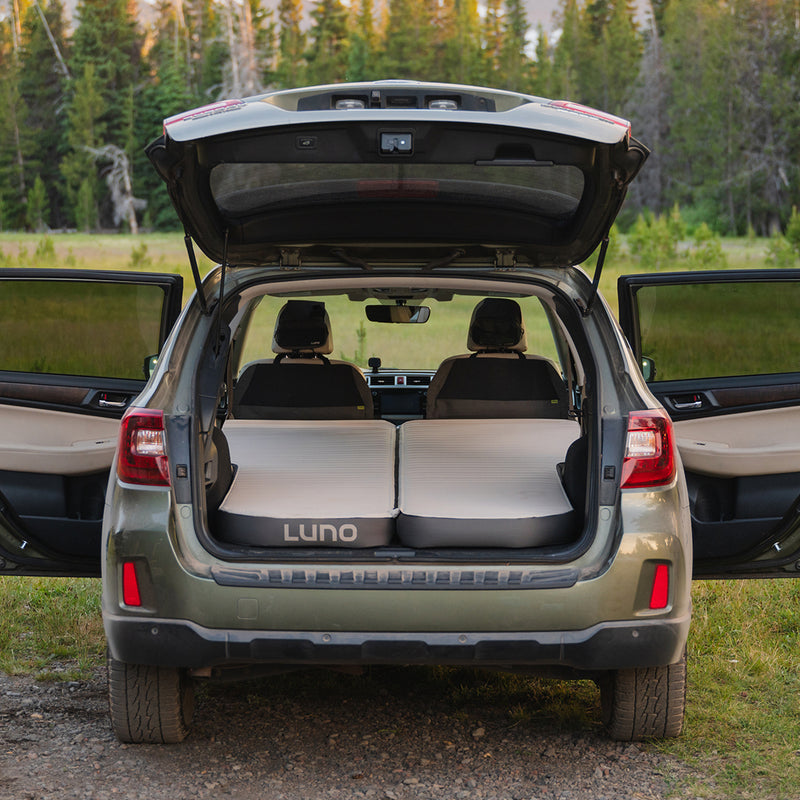 An SUV with its back hatch and rear doors open reveals the back seats folded down and two AIR+FOAM Pro Car Camping Mattresses set up inside for camping. A grassy field and trees are visible in the background.