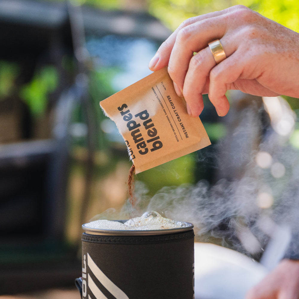 A hand adorned with a gold ring pours Luno® Campers Blend Instant Craft Coffee from a brown packet into a steaming black cup. The background is softly blurred, revealing greenery and hints of a car or tent, evoking the spirit of an outdoor adventure in a camping setting.