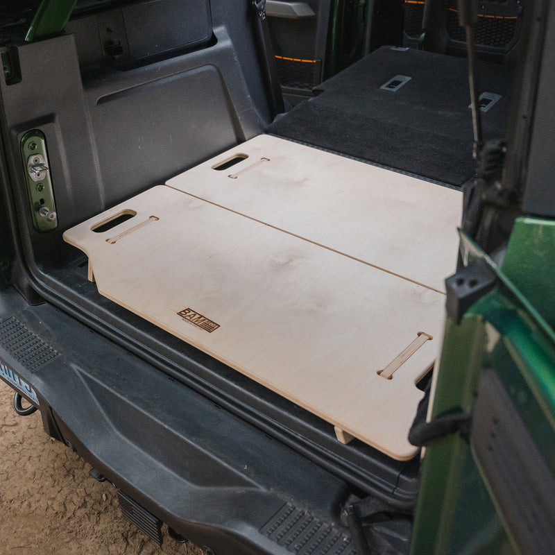 The Vehicle Sleeping Platform features two light wood panels with cut-out handles, installed flat in an SUV’s cargo area to create a modular bed over the rear storage space, with the back seats partially visible.