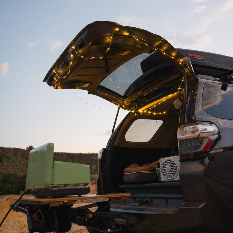 A dark SUV with its tailgate open, adorned with adjustable brightness Luno® Camping String Lights. Inside the back, there is camping gear and a green portable stove set up on a fold-out tray. A rechargeable USB-C battery pack powers the setup. The background features a serene outdoor landscape with a subtle evening sky.