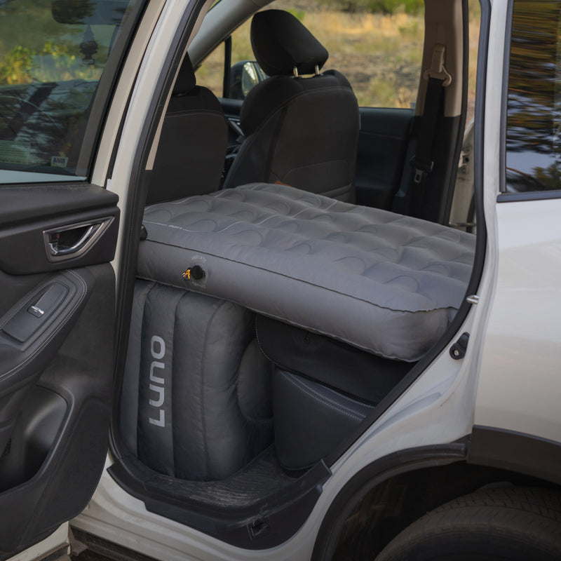 An AIR Vehicle Mattress (Single Side) by Luno HQ is set up in the back seat of a car, with the rear door open. It fits perfectly across the backseat, creating a cozy spot for solo camping. The vehicle is parked outdoors, suggesting an adventurous travel scene.