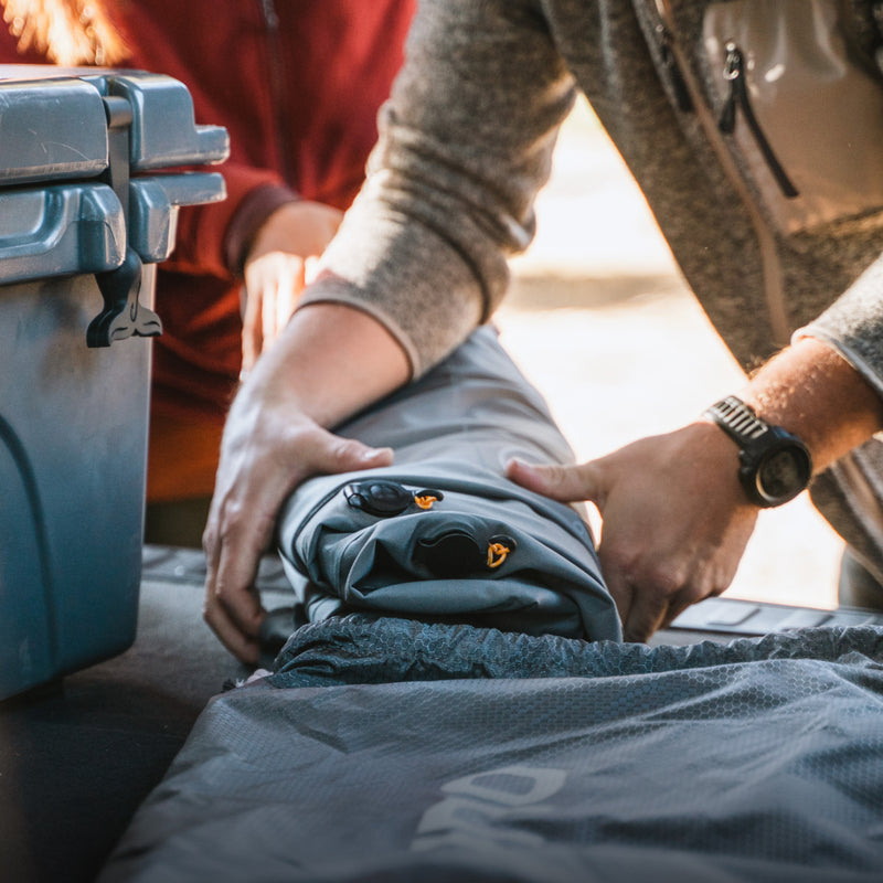 Two people are prepping gear for storage or travel. One rolls up a gray Luno HQ AIR Vehicle Mattress (Single Side) at the back of a vehicle, while another stands nearby. A blue storage container is visible, perfect for solo-camping adventures or storing upcycled items outdoors.