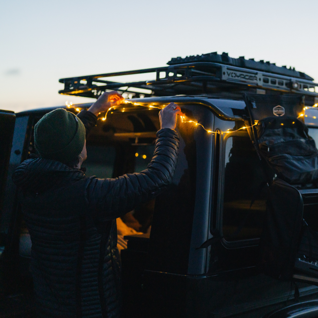 Wearing a beanie and puffer jacket, a person hangs Luno® Camping String Lights with adjustable brightness on a dark SUV at dusk. The lights, powered by a rechargeable USB-C battery pack, illuminate the roof rack and gear above.
