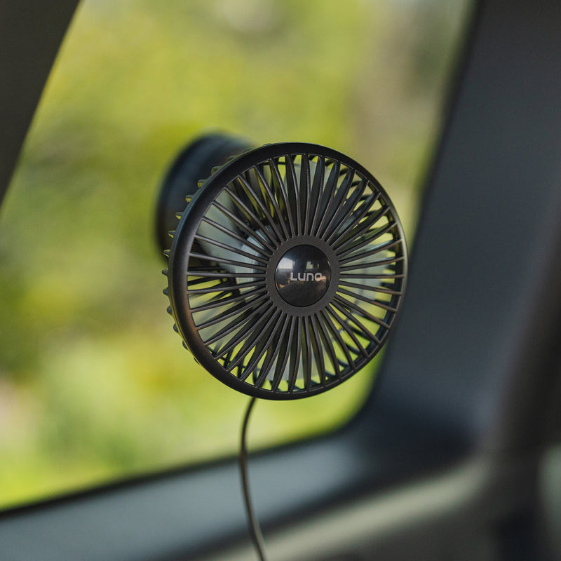 The Luno® Car Camping Cool Down Bundle includes a compact black camping fan with a circular grill, attached to a car window beside Car Window Screens—perfect for comfortable car camping with greenery outside. Powered by a visible cable.