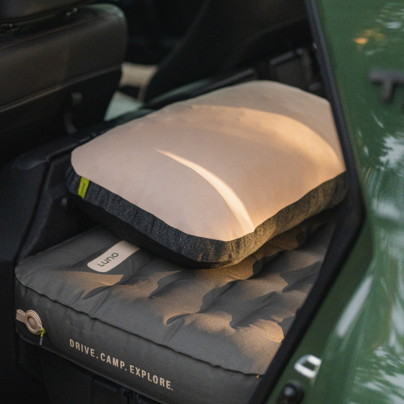 A car’s back seat is converted into a sleeping area with a gray inflatable mattress and Luno® Packable Camping Pillow. The mattress features “DRIVE. CAMP. EXPLORE.”.
