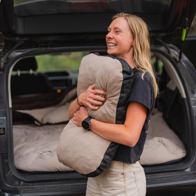 A smiling person stands by an open car trunk, hugging a tan and black sleeping bag, with Luno® Packable Camping Pillow and bedding visible inside the car.