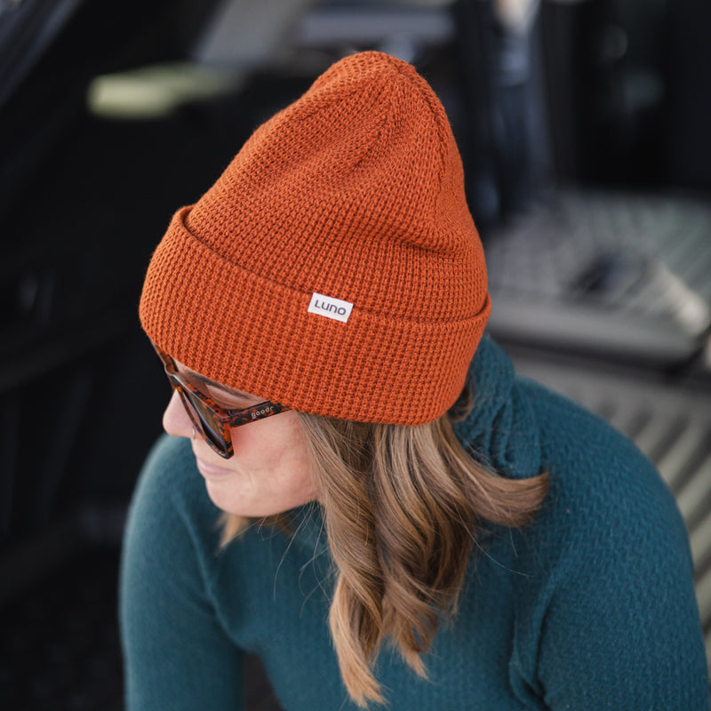 A person wearing a cozy burnt orange Waffle Knit Camp Beanie with a white Luno label and sunglasses sits in a car with an open trunk, paired with a teal sweater for a relaxed vibe.