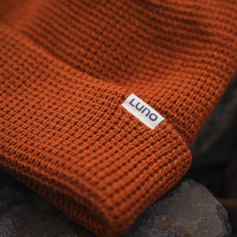 Close-up of a cozy burnt orange Waffle Knit Camp Beanie with a small white LUNO tag, resting on dark rocks.