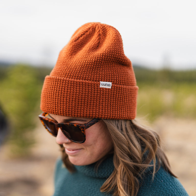 Wearing the Waffle Knit Camp Beanie in orange, tortoiseshell sunglasses, and a teal sweater, a person stands outdoors with a blurred natural background.