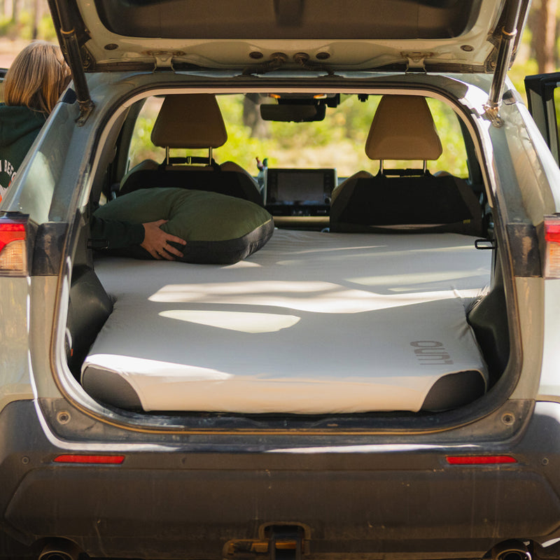 A person places a pillow inside the back of an SUV, where a Fitted Mattress Sheet - Vehicle Mattress covers an air mattress with seats down, preparing the area for camping or sleeping. Trees are visible outside through the open rear hatch.