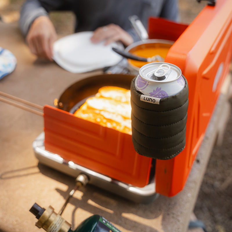 A Magnetic Can Cooler holding a Luna can is attached to an orange camping stove, with food cooking in a pan and a person holding a plate in the background.