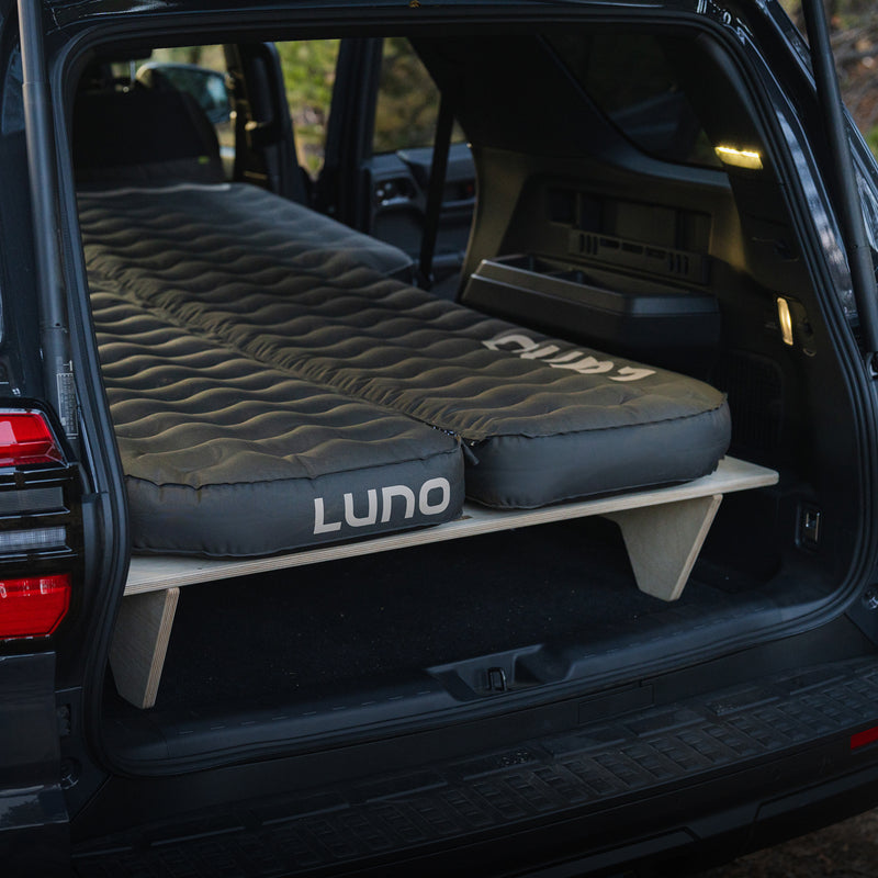 A black SUV with its rear hatch open displays a Vehicle Sleeping Platform supporting an inflated air mattress, set up for comfortable sleeping or camping inside the vehicle.