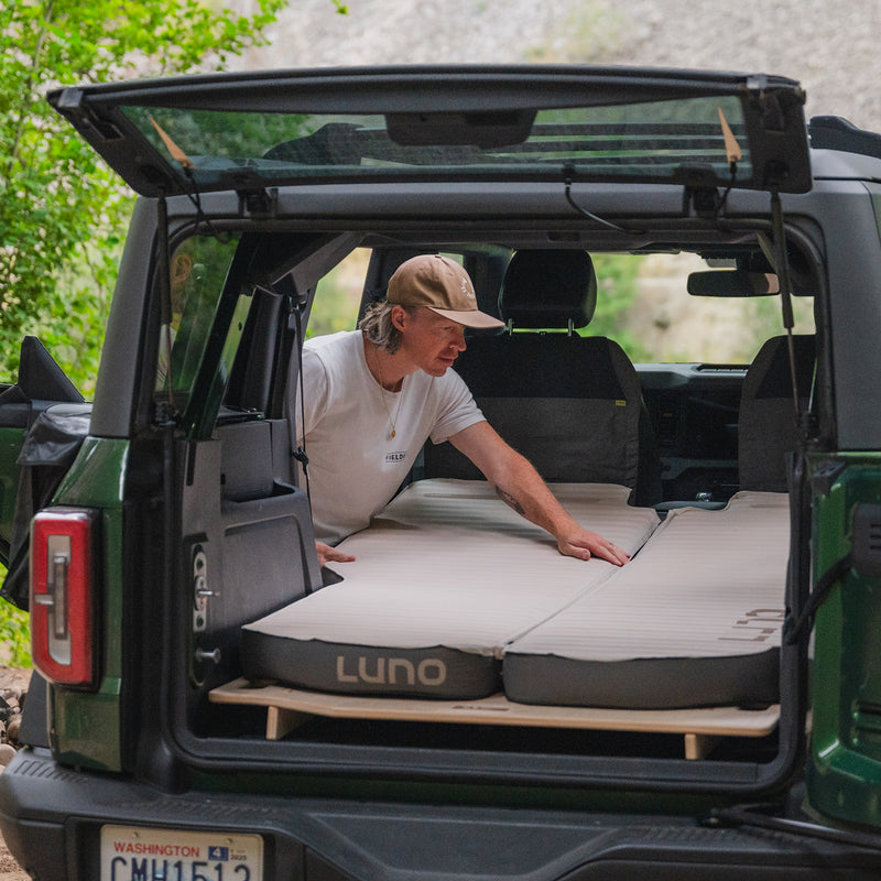 A person sets up a Vehicle Sleeping Platform in the back of an SUV, creating a comfortable camping space. The rear hatch is open, showcasing trees and greenery outside.