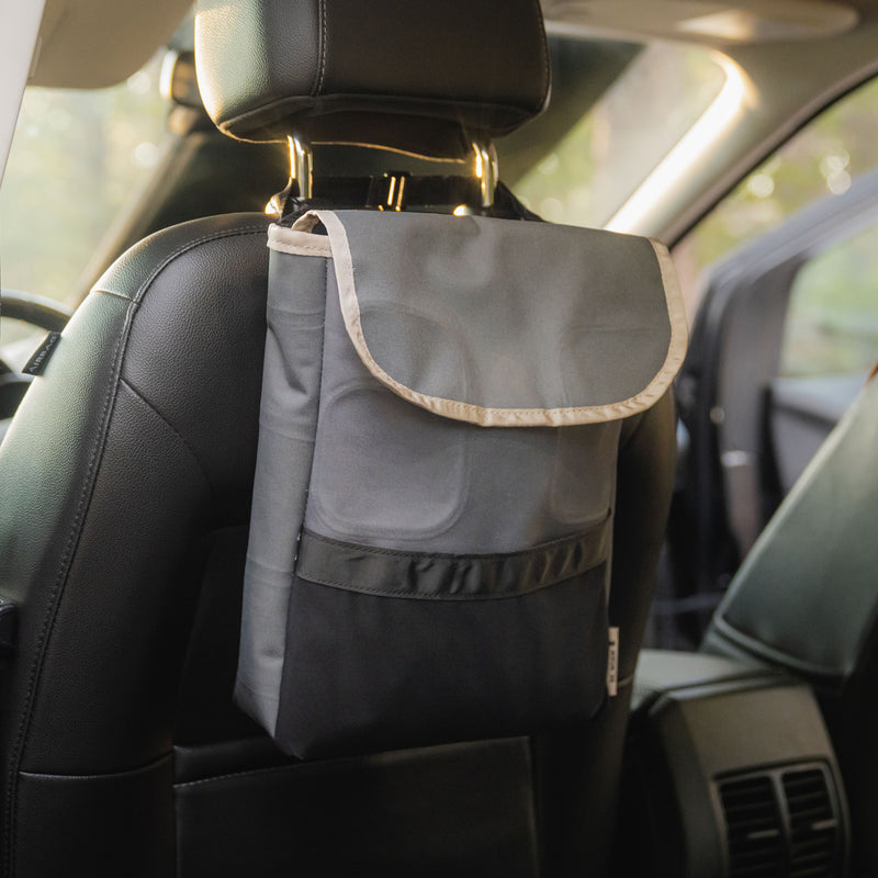 A Car Trash Can, designed in gray and black from upcycled materials, hangs from a car seat headrest inside a vehicle as sunlight filters through the windows in the background.
