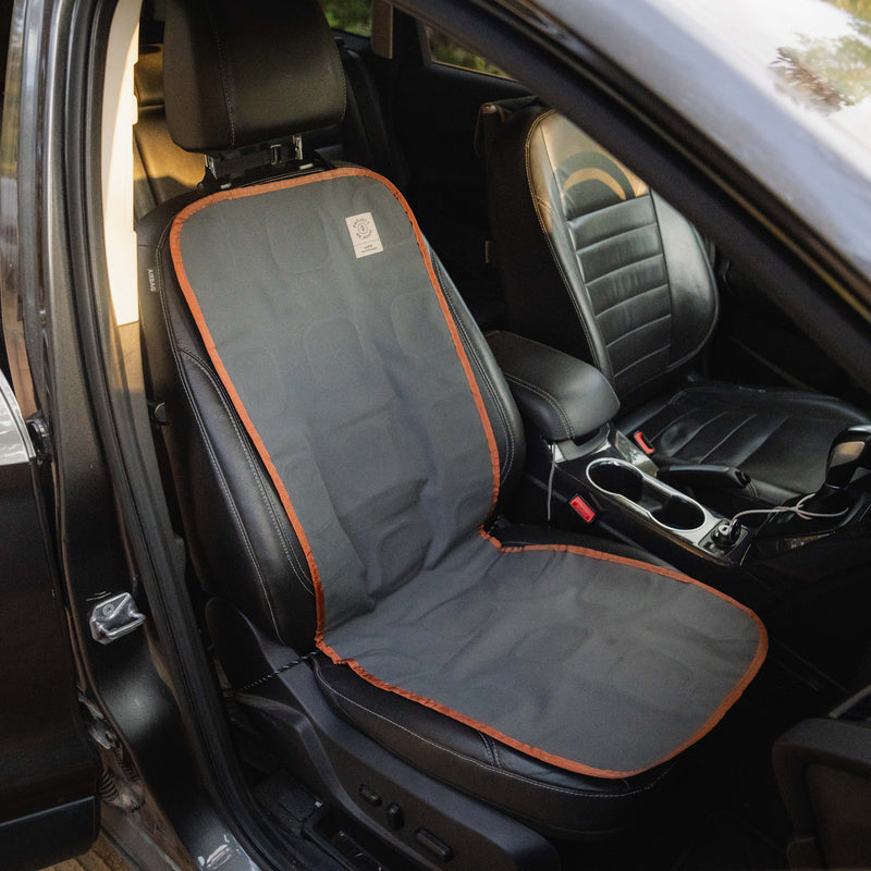 An All-Weather Seat Cover in fitted gray and orange is shown protecting a black car seat inside a vehicle, providing durable vehicle protection. The front passenger seat, center console, and part of the dashboard are also visible.