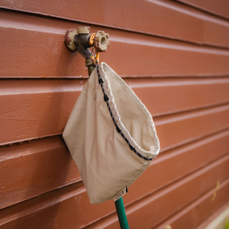 A Toiletry & Travel Organizer Pouch in beige, evoking the look of sustainable gear, is attached to an outdoor water spigot on a brown wooden wall for protection. A green garden hose is connected to the spigot and extends downward.
