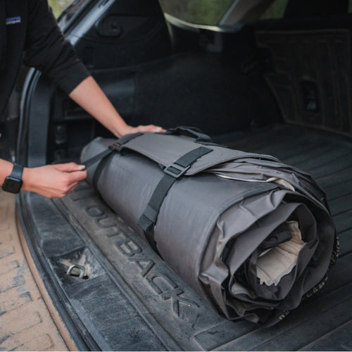 Person rolling up a camping mat and securing it with black straps inside the trunk of a car. The interior of the car is spacious and appears suited for outdoor gear storage. The person is wearing a black long-sleeved shirt and a smart watch on their left wrist.