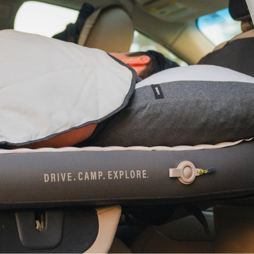 A person with short dark hair is lying on their stomach in the backseat of a car, resting their head on a gray pillow. They appear to be covered with a light-colored blanket. The text "DRIVE. CAMP. EXPLORE." is visible on a section of the car.
