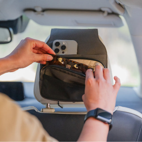 A person places sunglasses into a small storage compartment attached to the back of a car seat. An iPhone is secured in a dedicated slot on the storage organizer. The person is wearing an Apple Watch on their wrist.
