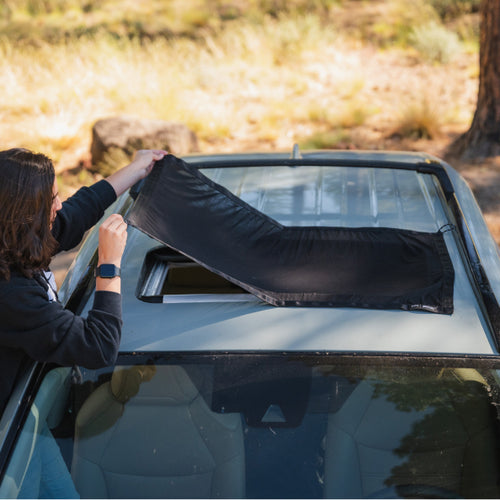 A person with long hair is attaching or removing a rectangular black mesh cover from the sunroof of a car. The surrounding area is outdoors, with trees and grass visible in the background. The person is wearing a black jacket and a smartwatch.