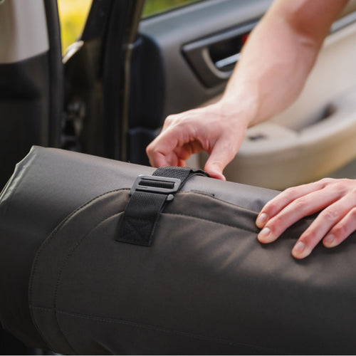 A person secures a rolled-up sleeping bag or mat with a black strap before placing it inside a car. The car door is open, and the interior of the vehicle is partially visible.