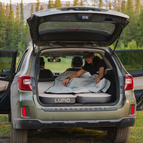 A person is setting up a sleeping area in the back of an SUV with the trunk open. They are placing a blanket over an inflated mattress labeled "LUNO". The scene is set in a scenic outdoor location with trees and mountains in the background.