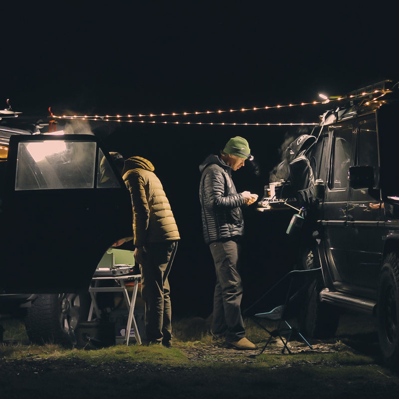 Two campers settle between two cars at night. One cooks on a portable stove, while the other checks the car. Luno® Camping String Lights hang between the vehicles, offering adjustable brightness and powered by a rechargeable USB-C battery pack, creating a cozy atmosphere.