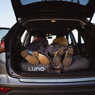 Two people lie on their backs inside the trunk of an SUV, with their legs extending outward. They rest on a Luno branded mattress, and the open trunk door reveals their relaxed, casual posture. The scene suggests a cozy camping or road trip experience.