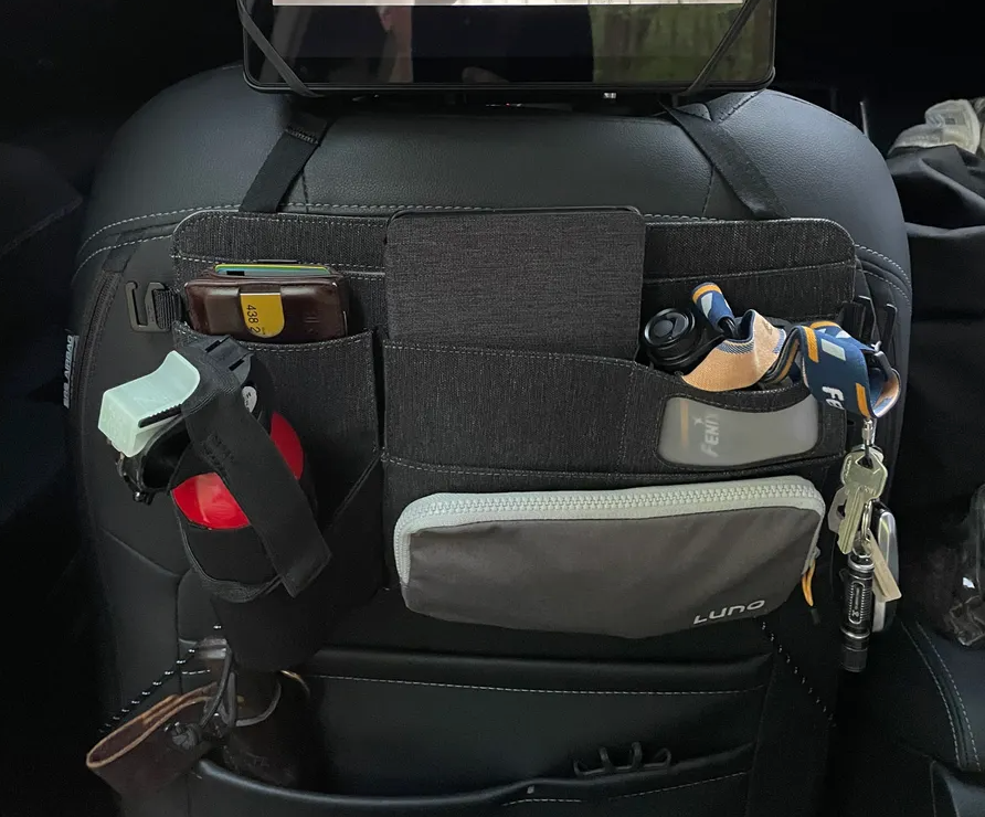 Car seat organizer attached to the back of a front seat, containing various items: a smartphone, tablet, flashlight, water bottle, keychain, and other small objects, neatly stored in different compartments and pockets.