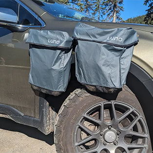 Two gray Luno branded storage bags attached to the exterior windows of a vehicle. The bags are mounted over the top of the car door windows, hanging on the side. The car is parked outdoors, with trees visible in the background.
