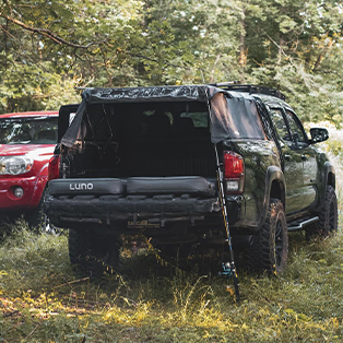 A black SUV with an open tailgate is parked in a wooded area next to a red vehicle. The SUV has a camping setup in its back, including an inflatable air mattress branded "Luno". The scene is surrounded by green foliage and appears to be a campsite.
