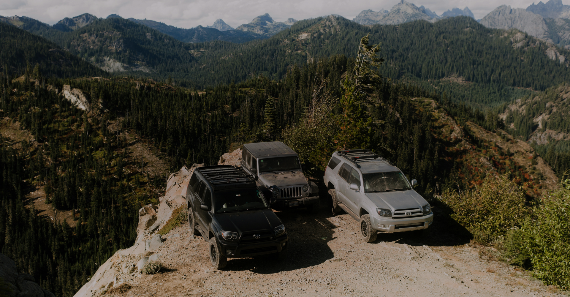 Three off-road vehicles are parked on a rocky overlook surrounded by a vast forest landscape with mountainous terrain in the background. The sky is partly cloudy, casting soft shadows over the picturesque scene, showcasing nature and adventure.