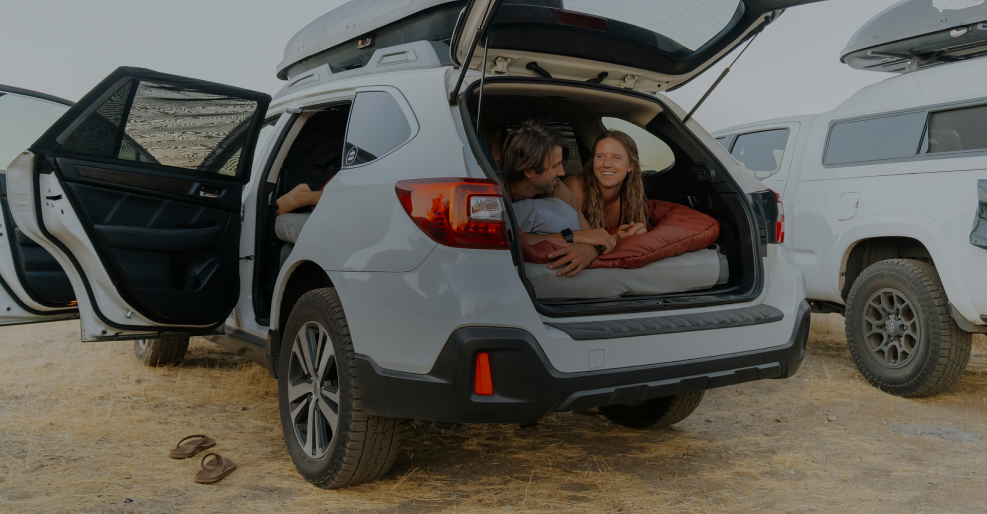 Two people lie in the back of a white SUV with the trunk open, smiling and enjoying a camping trip. Sandals are on the ground nearby, and another SUV is parked beside them. The outdoor setting appears sunny and relaxed.