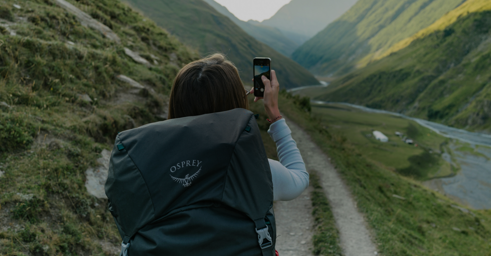 A person wearing a backpack stands on a narrow mountain trail, taking a photo of the scenic valley below with their smartphone. The valley features lush green slopes and a river winding through the landscape.
