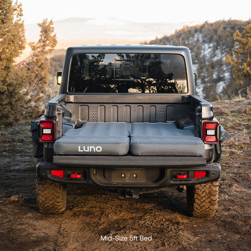 A pickup truck with its tailgate down features a Luno® AIR Truck Bed Mattress, creating a comfortable sleeping area across the 5-foot bed—ideal for camping. Trees and hills can be seen in the background.