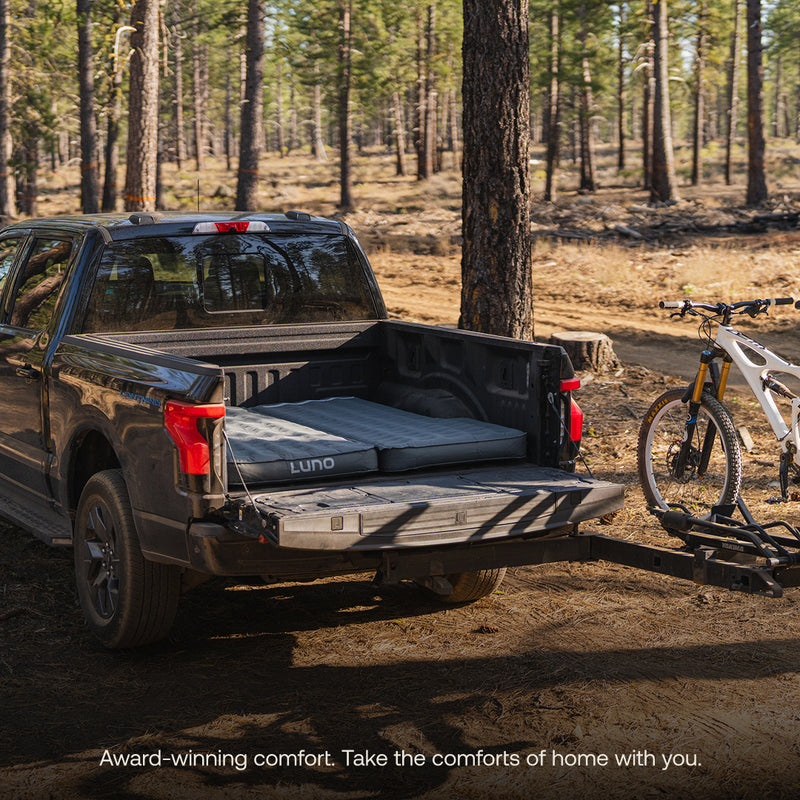 A pickup with a Luno® AIR Truck Bed Mattress in its bed is parked in a sunlit forest, mountain bike on the rear. Text: "Luno® Award-winning comfort. Bring home-like rest wherever adventure calls.