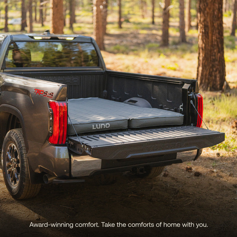 A pickup truck sits in a forest clearing with its tailgate down, featuring an optimized sleeping space created by the inflated Luno® AIR Truck Bed Mattress in the bed, ready for camping comfort.