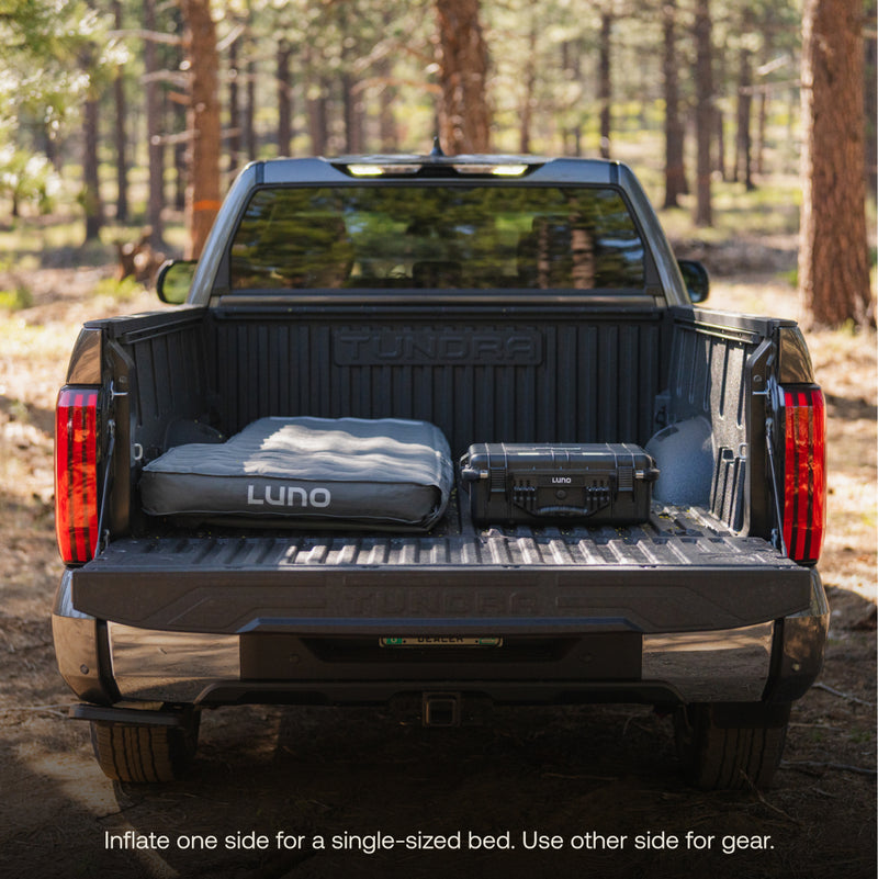 A pickup truck in the forest with its tailgate down. A Luno® AIR Truck Bed Mattress is partially inflated for a single bed, while the other side stores gear. Text: "Inflate one side for sleeping, use other side for storage.