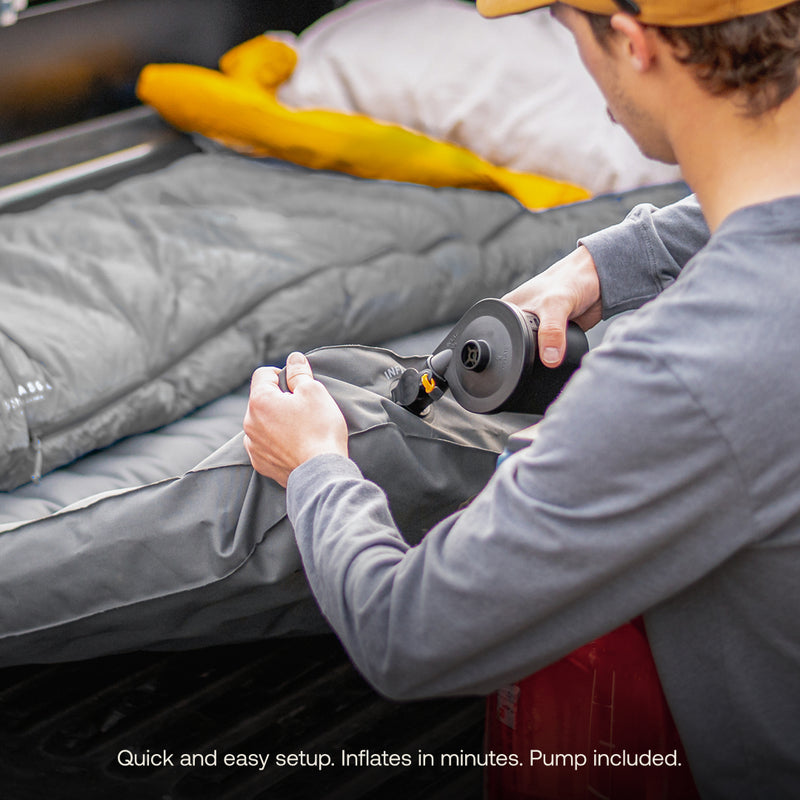 A person uses a pump to inflate a gray Luno® AIR Truck Bed Mattress in a vehicle's bed. A sleeping bag and yellow pillow are nearby. Text: "Quick, easy setup. Optimized sleeping area. Pump included.