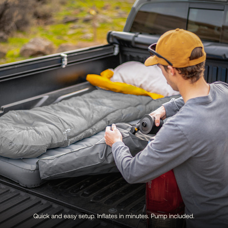 A person in a tan cap uses the included pump to inflate a Luno® AIR Truck Bed Mattress with a gray sleeping bag outdoors. Text: "Quick, easy setup—inflate in minutes with included pump for a comfortable sleeping area.